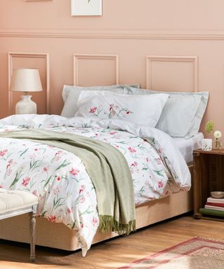 A peach pink bedroom with white pink tulip patterned bedding with pillows and a duvet, a sage green throw over the bed, and a white tablelamp to the left and a dark wooden nightstand to the right