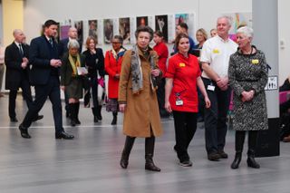 Princess Anne wearing a tan coat and black boots walking through a hospital with staff members next to and behind her