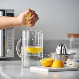 Jug of water with a lemon half in it, next to a microwave and halved lemons on a chopping board beside it