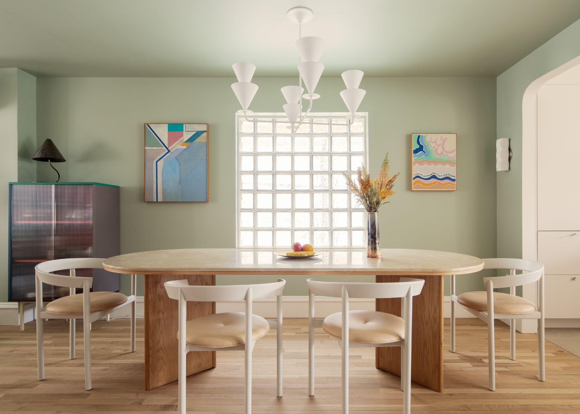 A modern open-plan dining room with sage green color-drenched walls, wooden flooring, a large curved dining table with modern white chairs, and a glass brick wall.