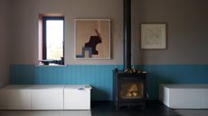 Image of a wall that is painted gray with bright blue paneling across the bottom half of the wall. There is low bench seating with storage and a black, old-fashioned fireplace. 