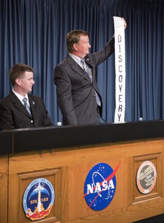 Space Shuttle Program Launch Integration Manager Mike Moses looks on proudly as Shuttle Launch Director Mike Leinbach holds up a Discovery banner signed by the STS-133 astronauts, at a news conference held in the Press Site auditorium at NASA's Kennedy Sp
