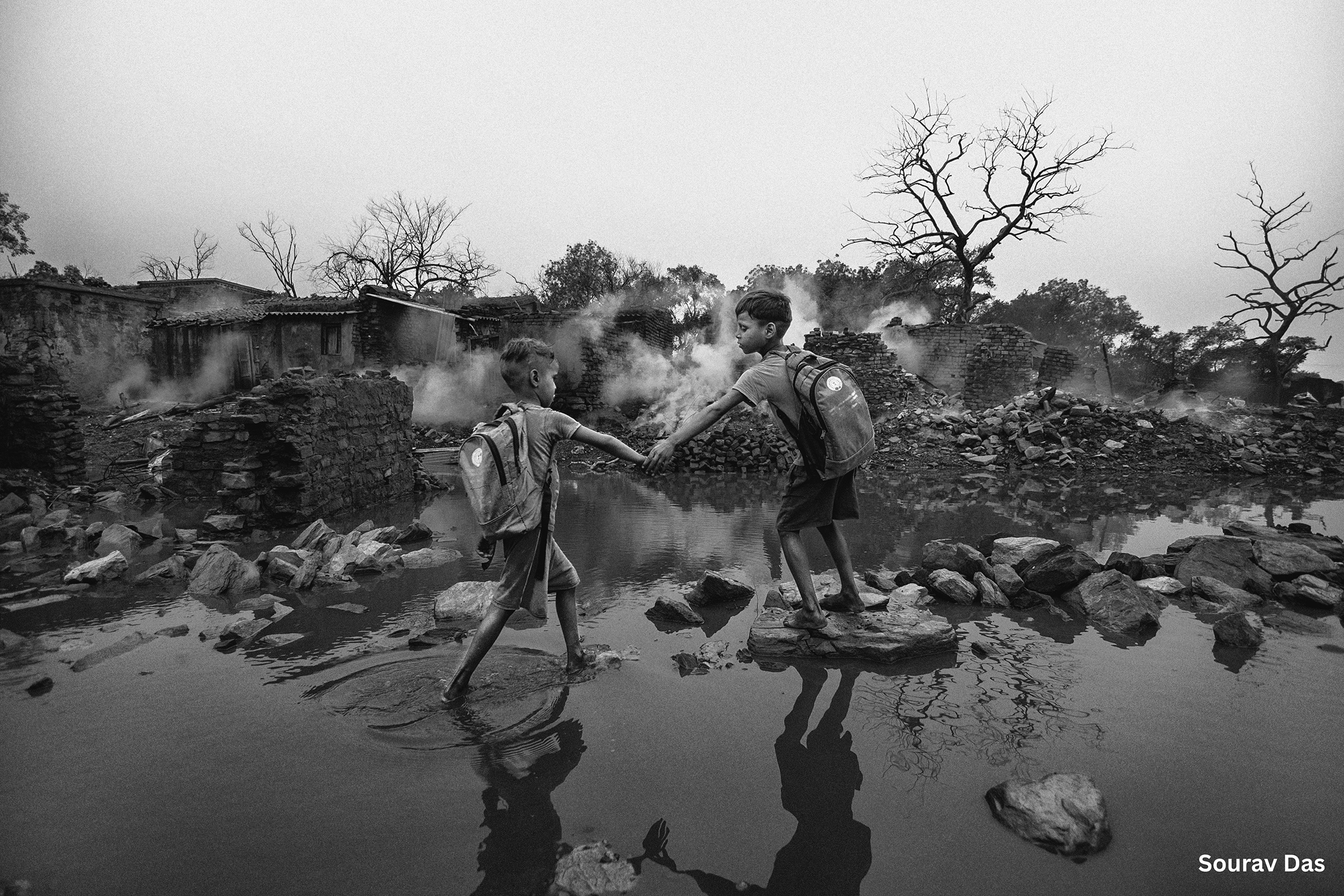 Two boys with backpacks hold hands while crossing a rocky, flooded area. Bare trees and ruins in the background create a somber and resilient mood
