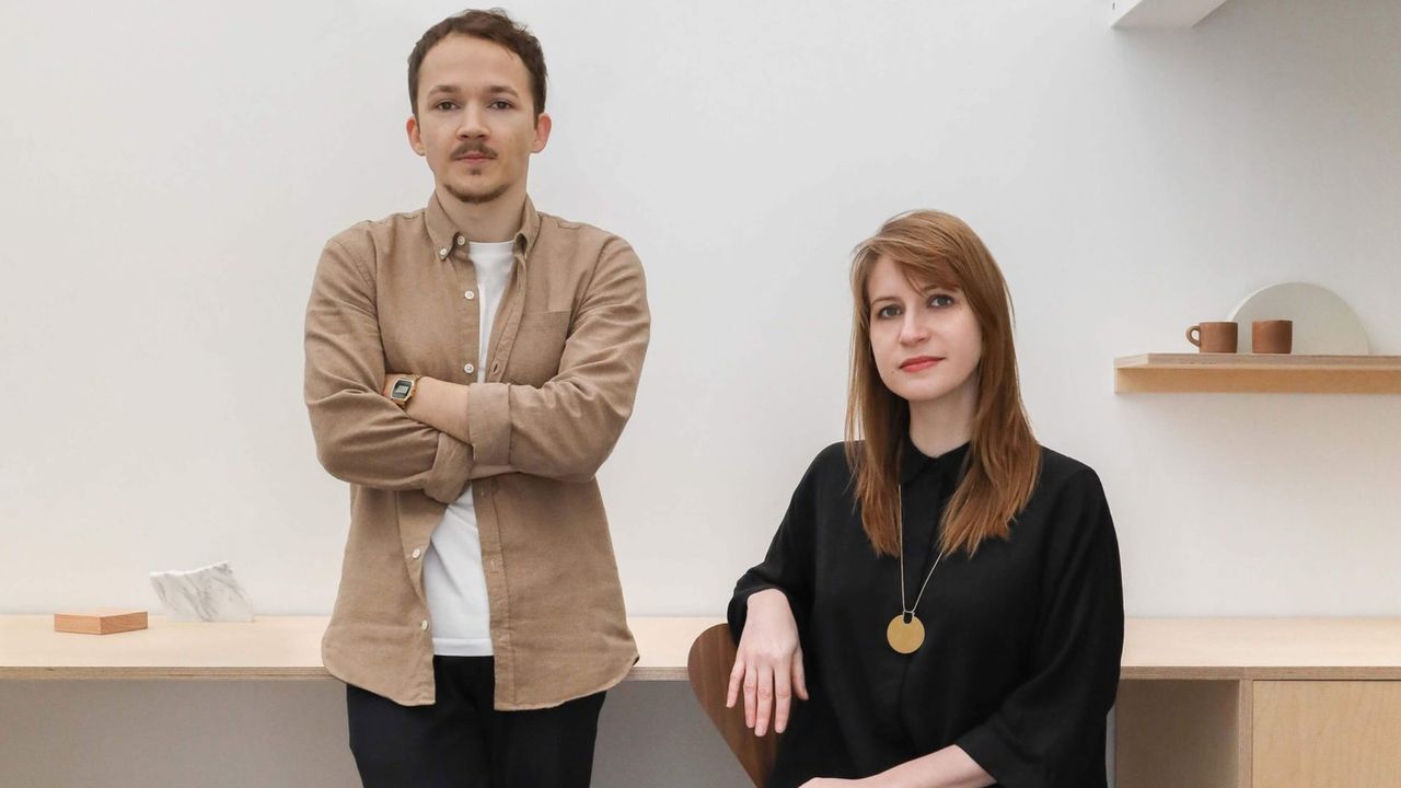 A young man dressed in a brown overshirt, white T-shirt, and black trousers and a young woman wearing a long-sleeved, black top stare at the camera in their simply decorated design studio.