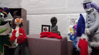 A fossilised Ara sits motionless with his friend surrounding him at a table, his work on Silksong news done.