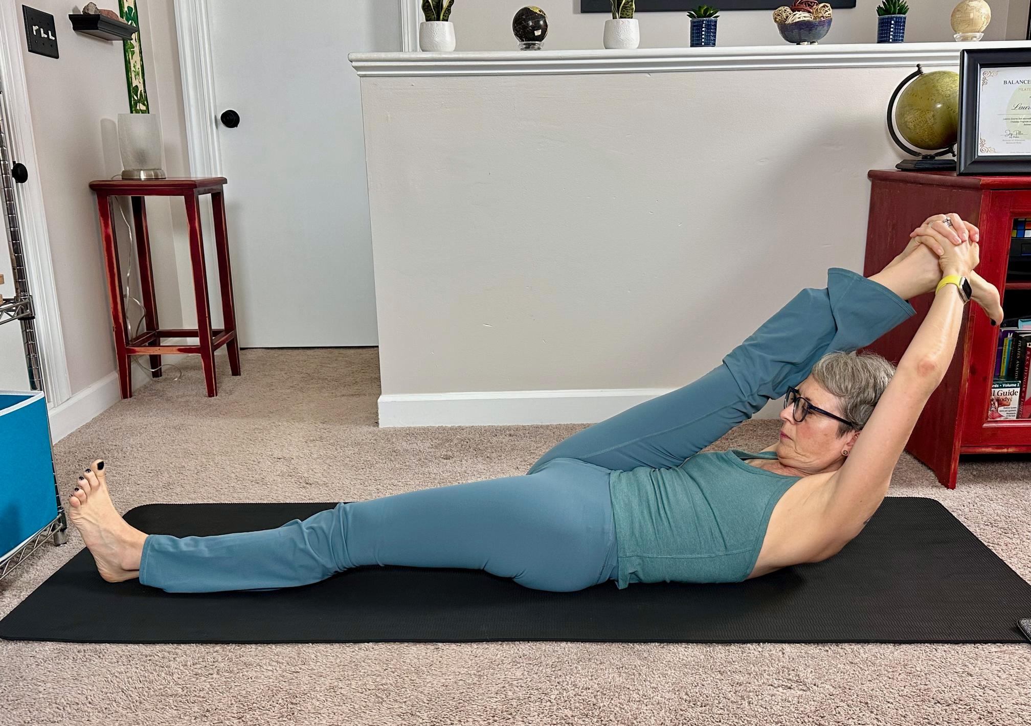 woman lying on her back on a black yoga mat in a living room performing a leg stretch with a straight leg extended toward her chest