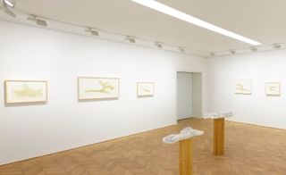 White room, wooden floor, white spotlights on a frame around the edge of the ceiling, two wooden posts topped with white model exhibition piece, framed pictures in a row around the centre of the gallery walls