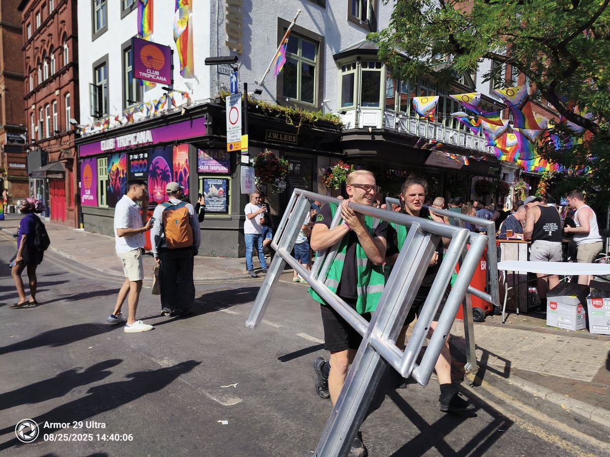 Two people in high-visibility vests carry a large metal barrier down a street decorated with rainbow flags and banners for "Club Tropicana."
