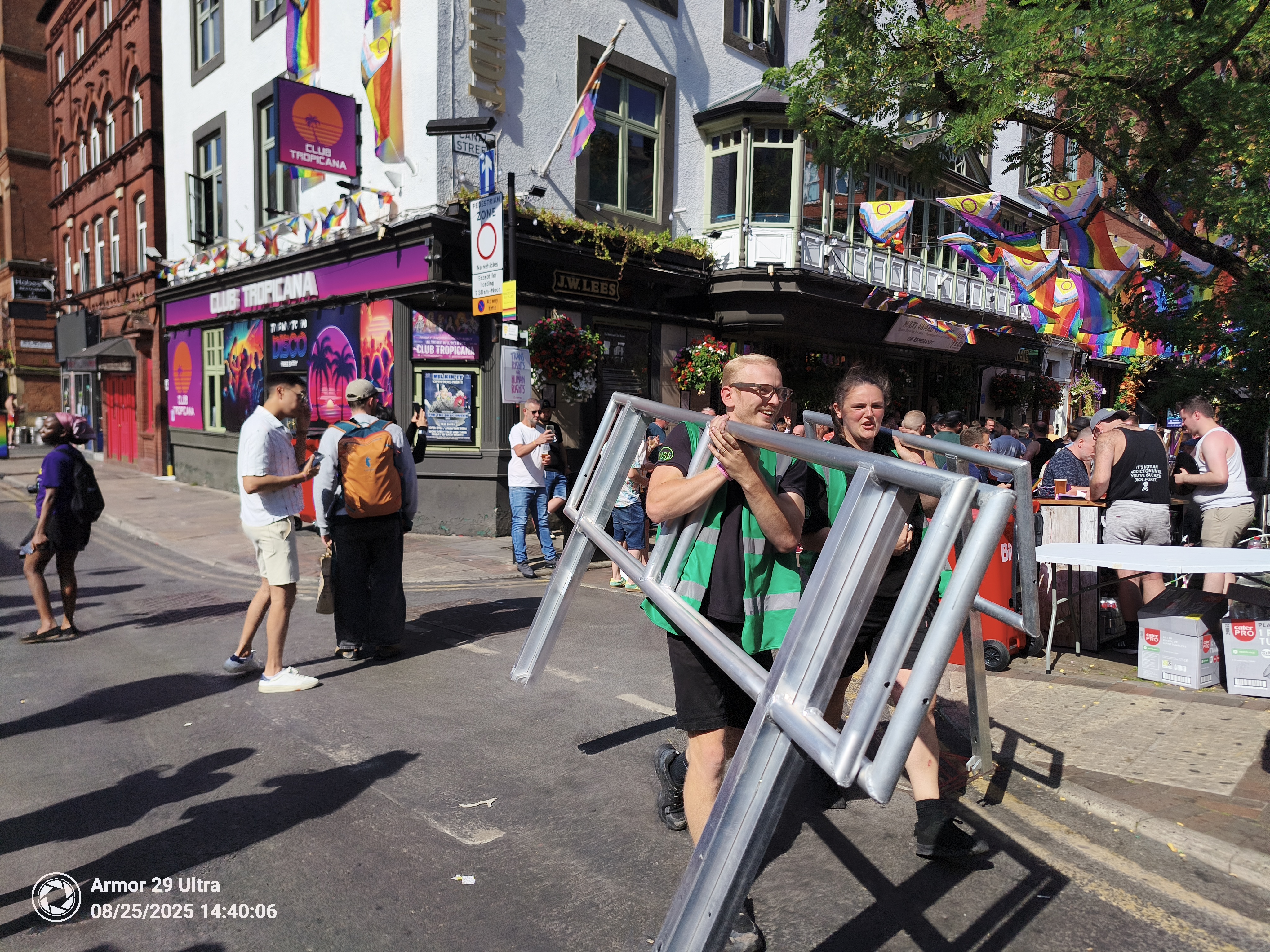 Two people in high-visibility vests carry a large metal barrier down a street decorated with rainbow flags and banners for "Club Tropicana."