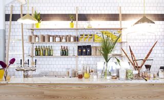 White tiled wall with bottles and coffee standing on shelves and a white counter top with coffee and a glass vase with greenery inside