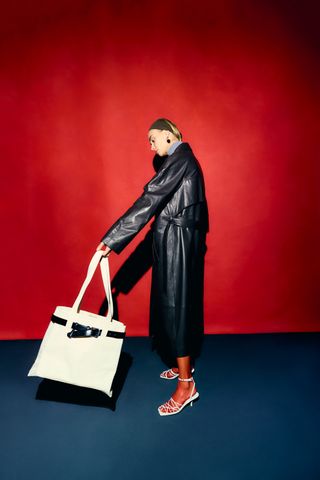 a model wears a leather trench coat while swinging llbean's tote collaboration with tibi in front of a blank wall