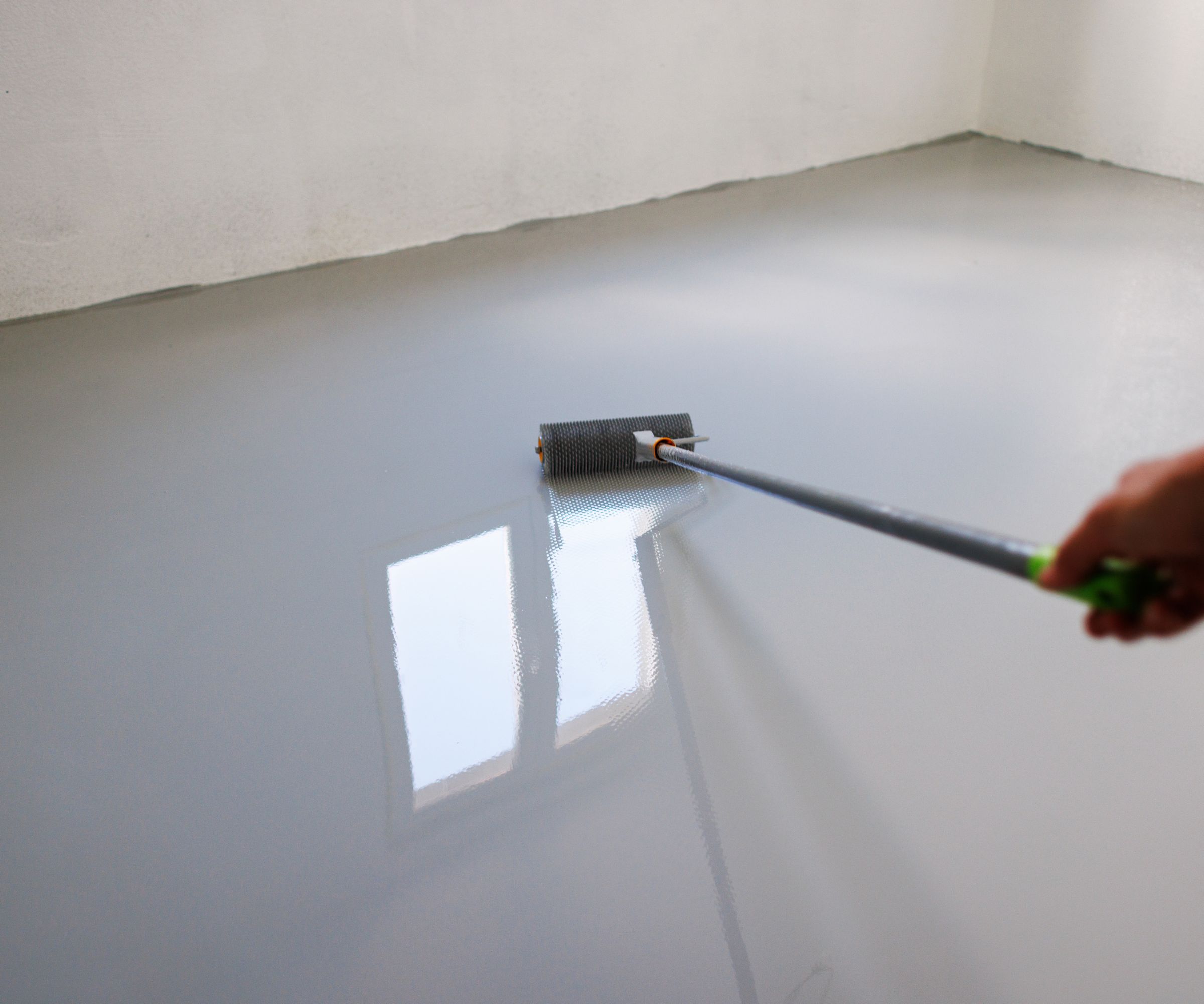 A close-up of a hand using a spiked roller to apply epoxy resin coating on a newly leveled floor. The reflective surface highlights the professional finishing process in a bright, natural-light environment.