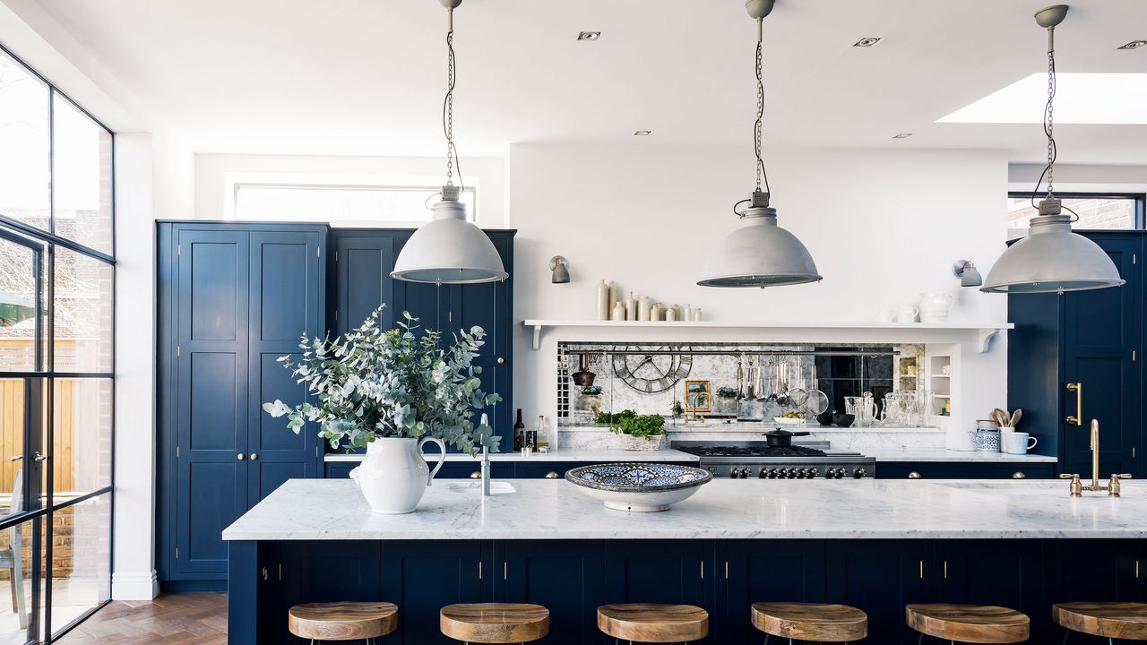 How to paint kitchen cabinets, illustrated in a white kitchen scheme with dark blue cabinetry and pendant lights over a marble breakfast island.