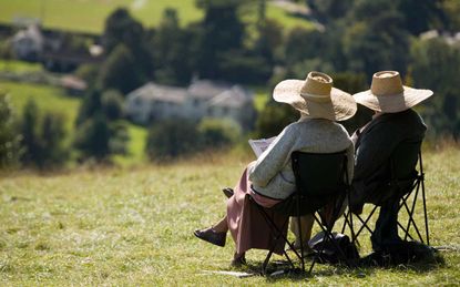 A pair of retirees sit on foldable lawn chairs atop a grassy hill in a pleasant town.