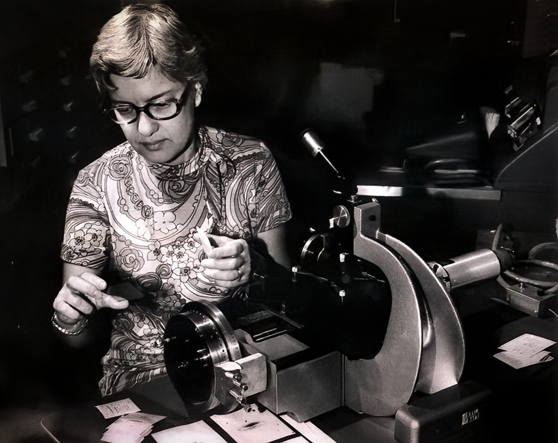 A woman with glasses sits next to a machine in a black and white photograph