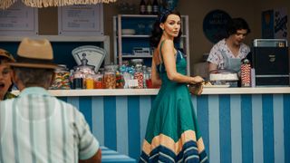 Michelle Keegan in character as nurse Kate Thorne wearing a green strappy dress and standing at a coffee counter