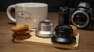 A black and a silver Laowa 15mm f/5 Cookie FF lens, on a board surrounded by cookies, a Nikon camera and a mug