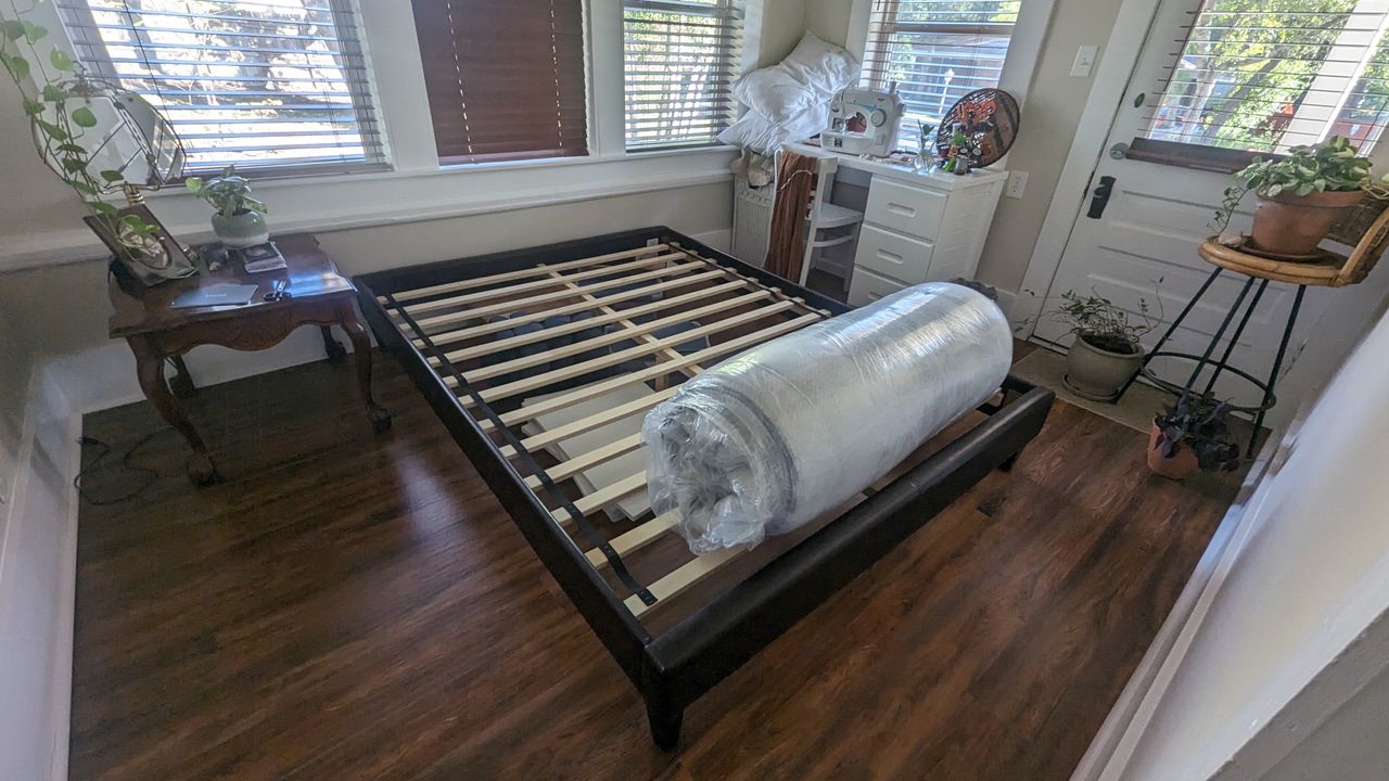 Review image shows the Leesa Oasis Chill Hybrid mattress rolled in plastic and sat on our tester's black bedframe waiting to be opened