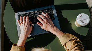 Woman typing on a laptop sitting down at a table next to coffee