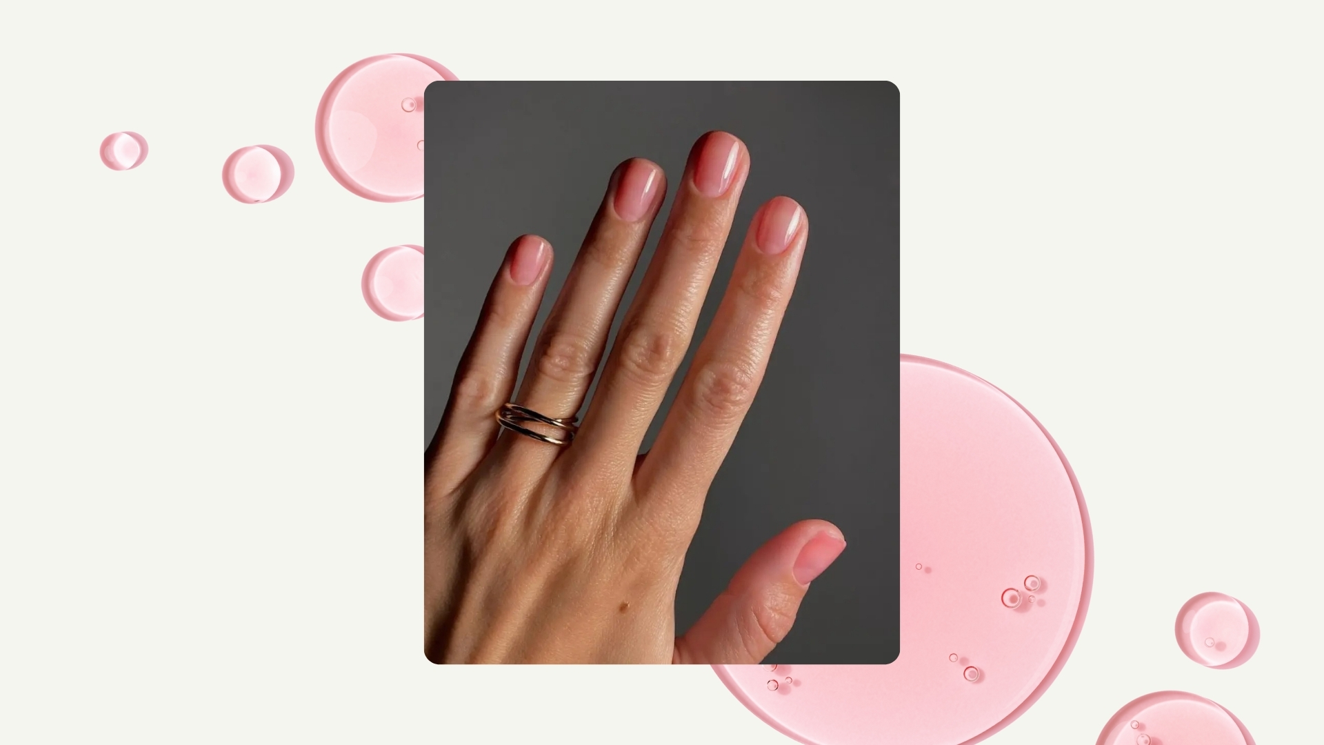 A close-up of a hand with sheer pink 'Rosehip oil'-like nails, by Digital Content Creator and nail artist, Mateja Novakovic/@matejanova, pictured in front of a grey background and in a cream template with pink oil drops