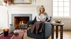 Designer Shea McGee photographed for her Target 2025 collection sat on a navy slipcovered armchair beside a fireplace with a wooden turned side table and a lacquered red coffee table