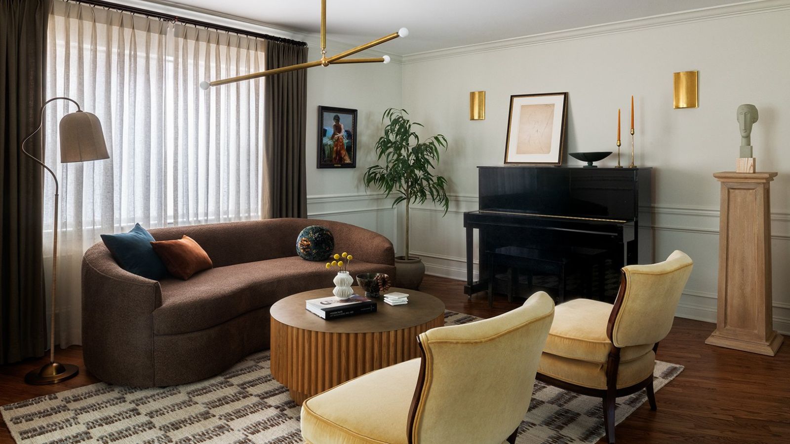 Living room with curved, brown, velvet sofa; round, brown coffee table; two cream armchairs; a black piano; an abstract, golden light hanging from ceiling; and ornaments pictured to the left and right.