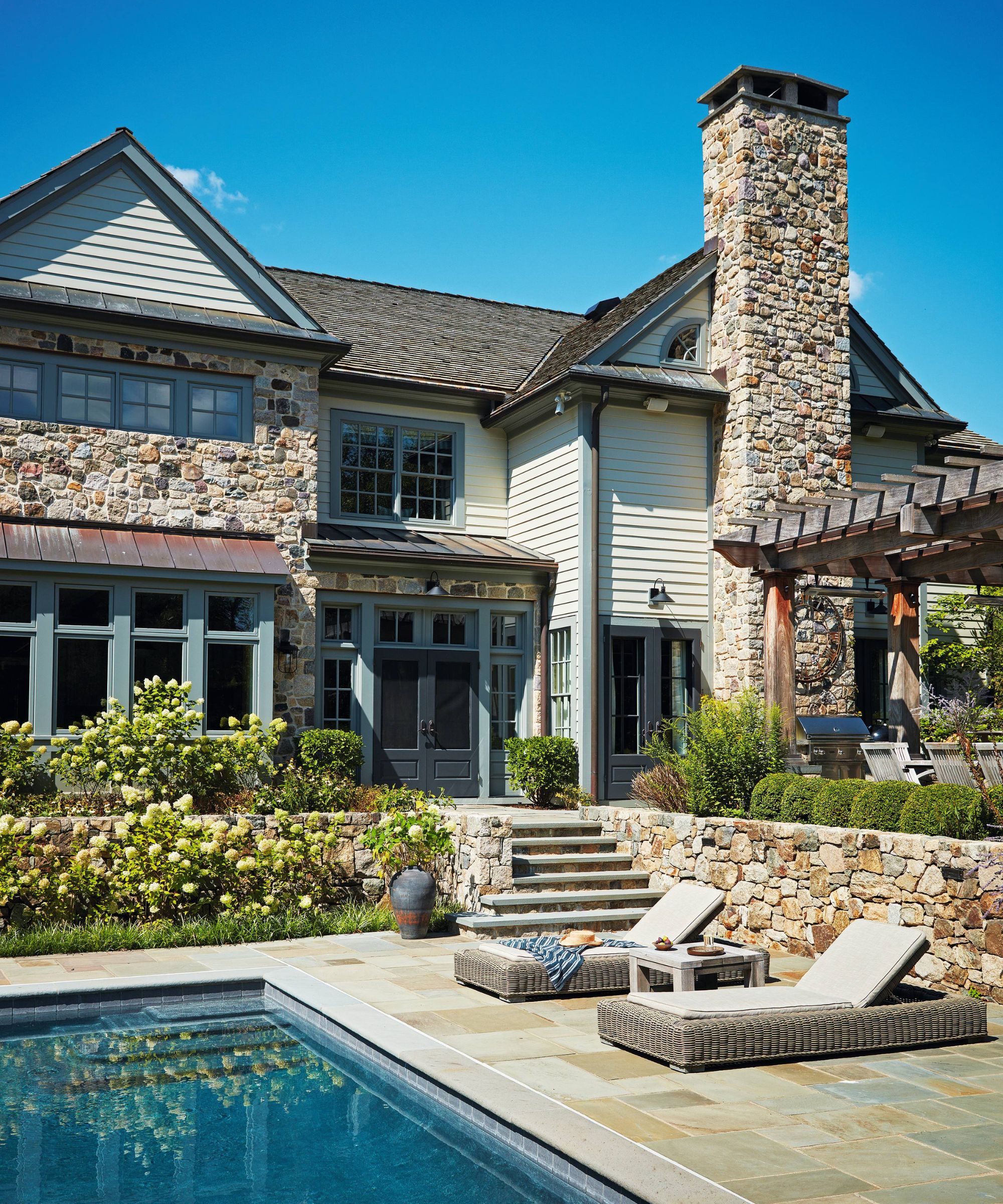 a large New Jersey colonial home with stone chimney and walls leading into a pool area with rows of hydrangeas
