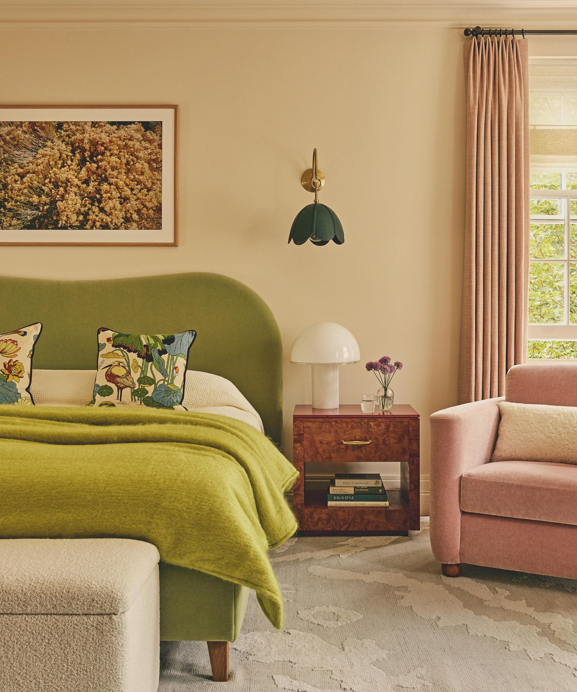 a soft pink bedroom with pink drapes and matching armchair with a green velvet bed, burl wood nightstand and flower shaped sconce