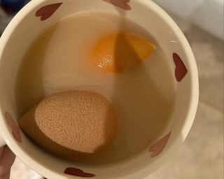 Two orange makeup sponges in dirty water in a white patterned mug