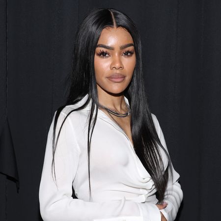 Teyanna Taylor standing in front of a black backdrop wearing a white shirt and necklace. She has long straight hair. 