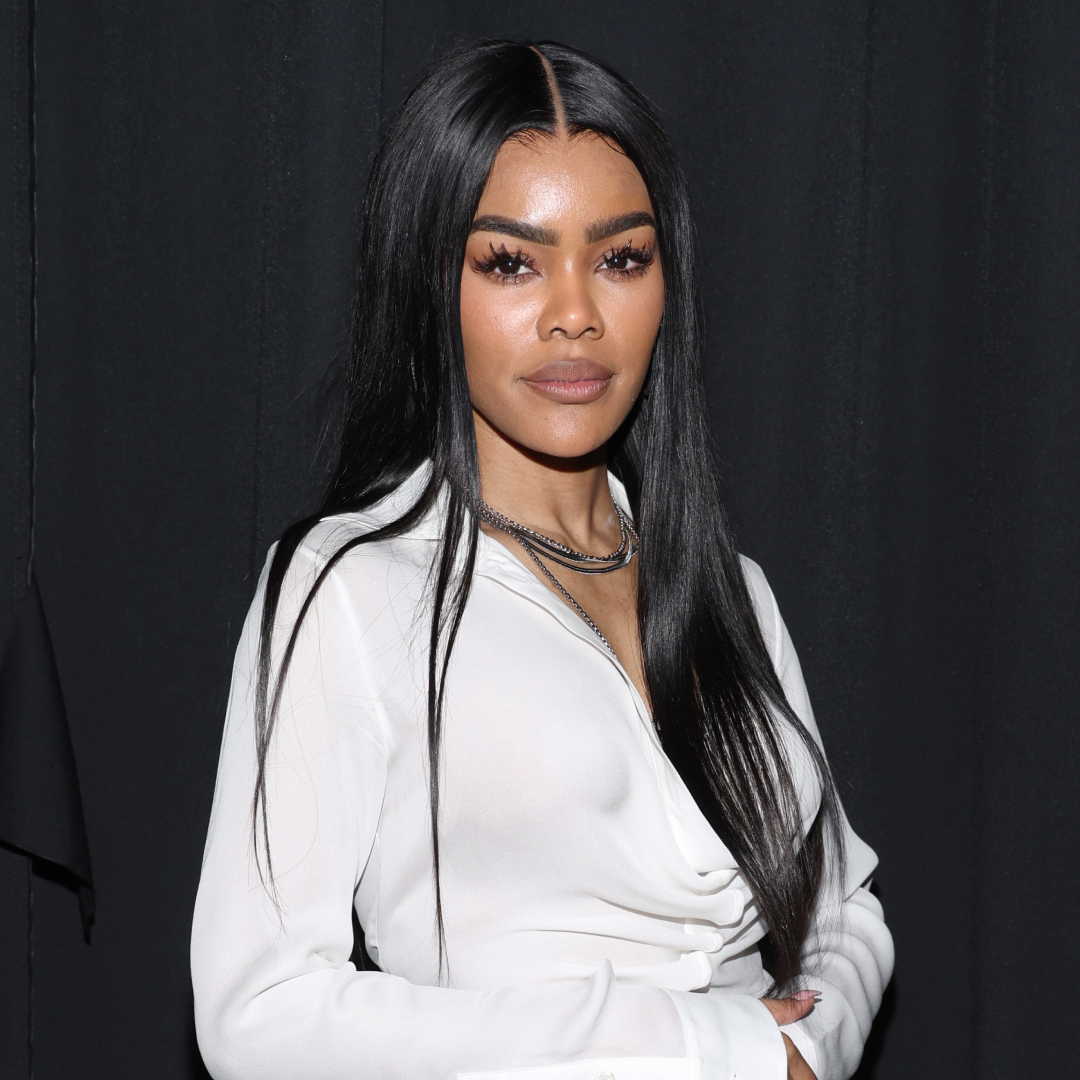 Teyanna Taylor standing in front of a black backdrop wearing a white shirt and necklace. She has long straight hair. 