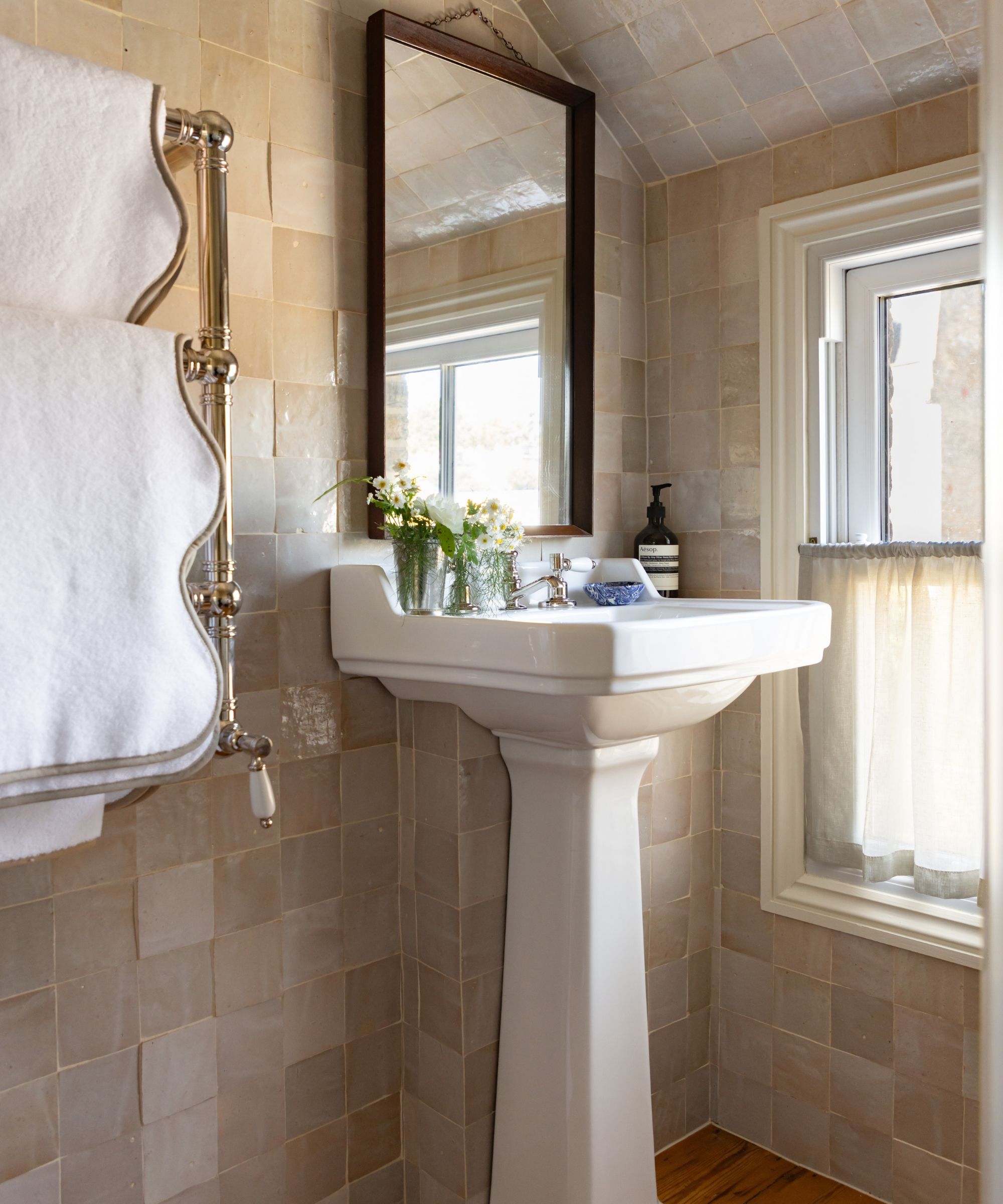 A bright bathroom with pale cream zellige tiles, a white sink, dark wood mirror, and towel rail with scalloped white towels. To the right is a window with a white cafe curtain.