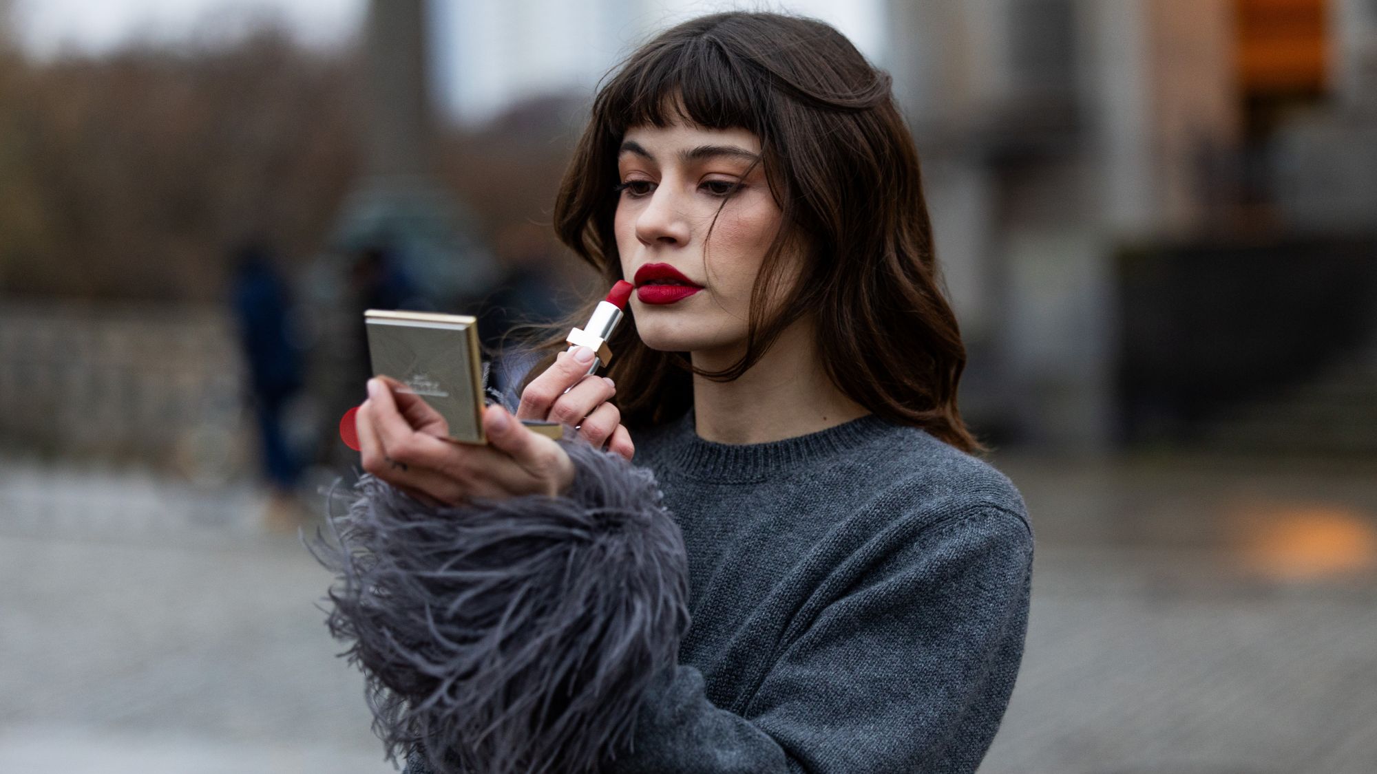 street style shot of woman in grey jumper applying red lipstick