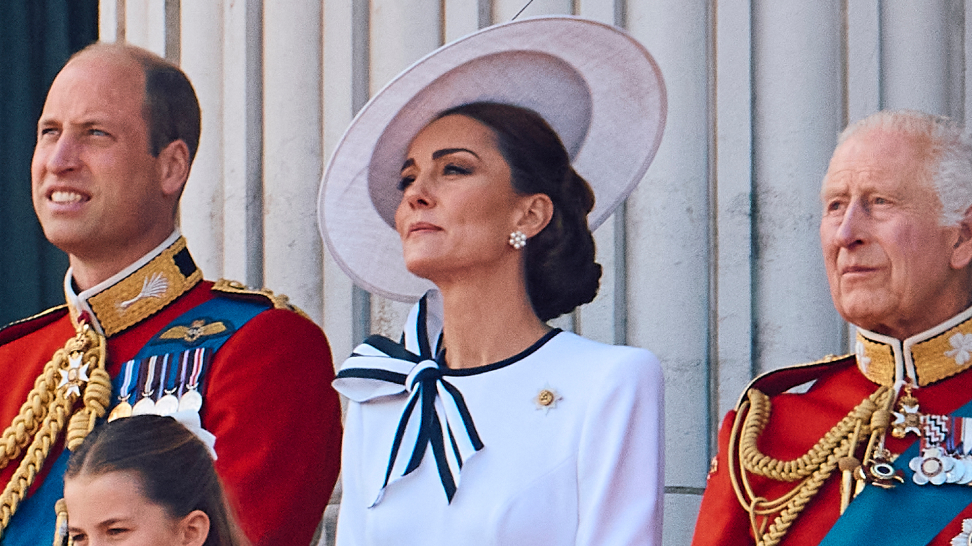 Prince William and King Charles wear red military uniform and Kate Middleton wears a white dress with navy accents and a matching hat on the Buckingham Palace balcony