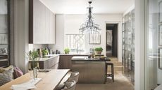 A neutral kitchen with chandelier, island, black bar stools, house plants, and a wooden kitchen table in the foreground.