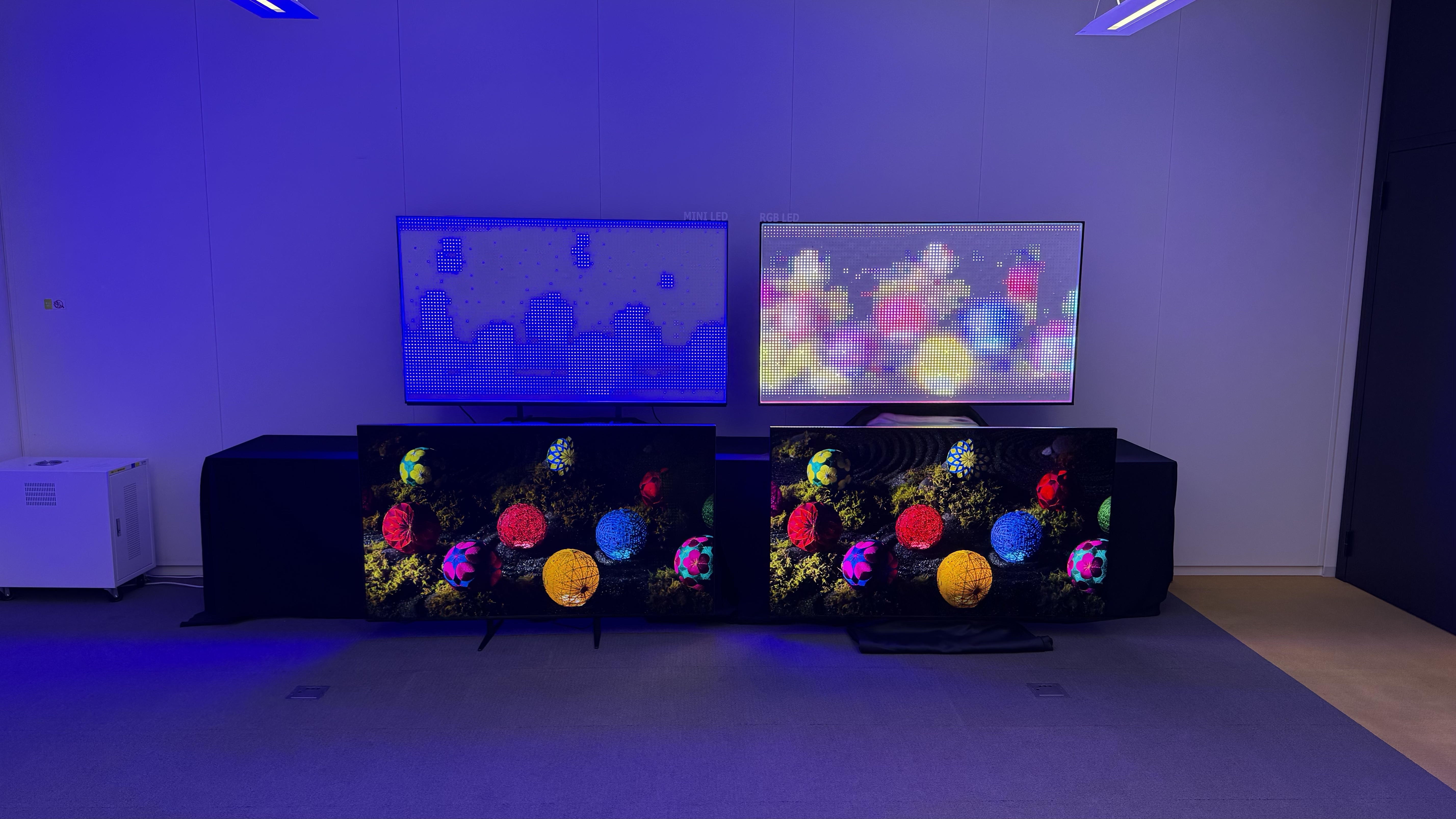 Two TVs arranged next to one another on the floor, with the exposed backlights of each on a table above. On both of the assembled TVs, an image of coloured lanterns is displayed. On the left-side backlight, a vague blue image is visible. On the right-side backlight, the colours are full recreated and the shapes of the lanterns are clearer.