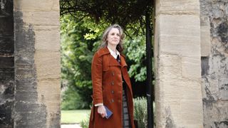 Abigail Thaw in a brown coat and checked trousers stands in an archway carrying a notebook as Dorothea Frazil in Endeavour.