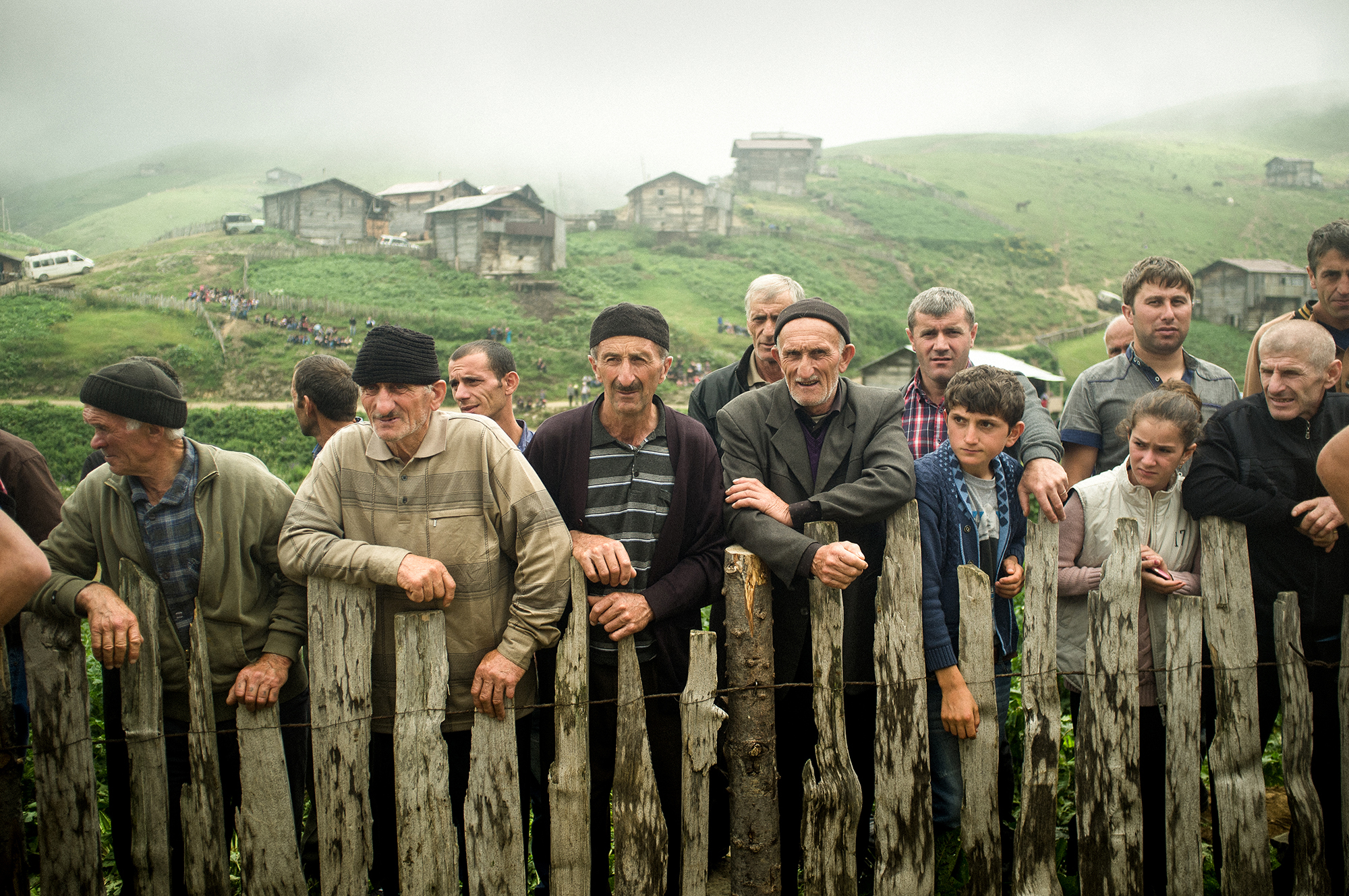 A group of people stand outdoors behind a rustic wooden fence on a grassy, foggy hillside.