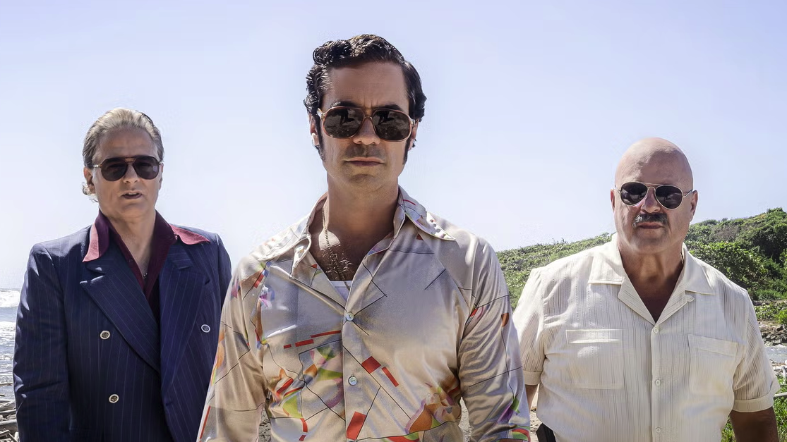 Cuban exile Roman Compte (Danny Pino) pictures between two henchmen on a sunny hillside wearing 1970s clothing in crime drama Hotel Cocaine 2024