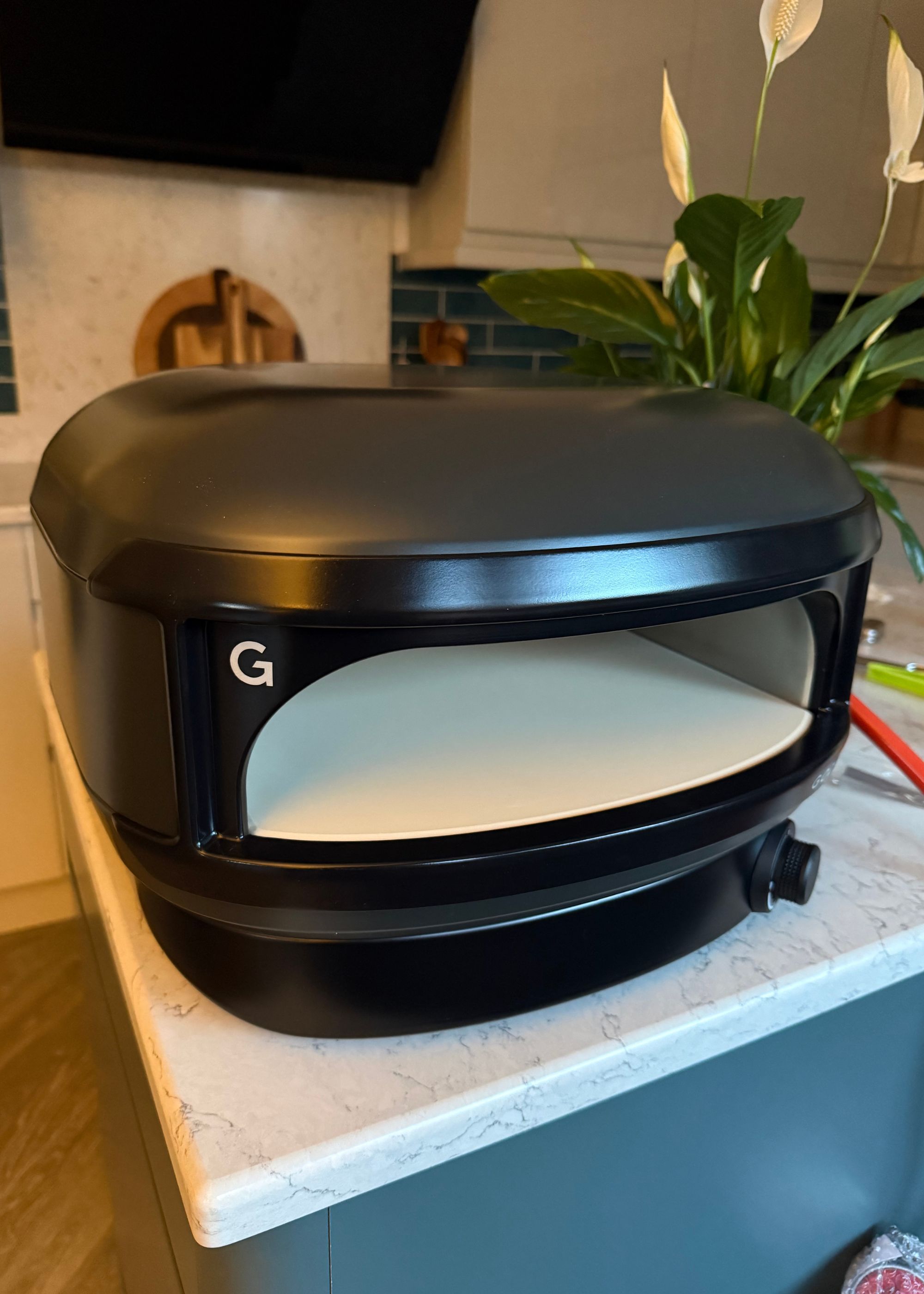 Gozney's ARc Lite pizza oven in off black sitting on top of a kitchen island next to a peacy lily plant with chopping boards and an extractor fan in the background next to the cabinetry