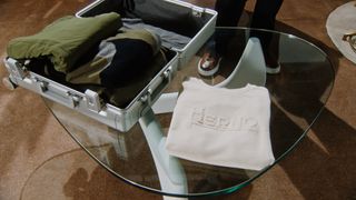 Open silver suitcase with folded clothing inside, on a glass curved triangular shaped coffee table on grey legs, folded cream sweater, rustic brown carpet, person in brown and white edged trainers and black pants in the back shot