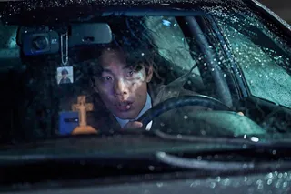 A man (Ryu Jun-yeol) looks out of a shattered car windshield while sitting behind the wheel, with a phone, wooden cross, and picture keychain visible, in the Netflix film 'Revelations.'