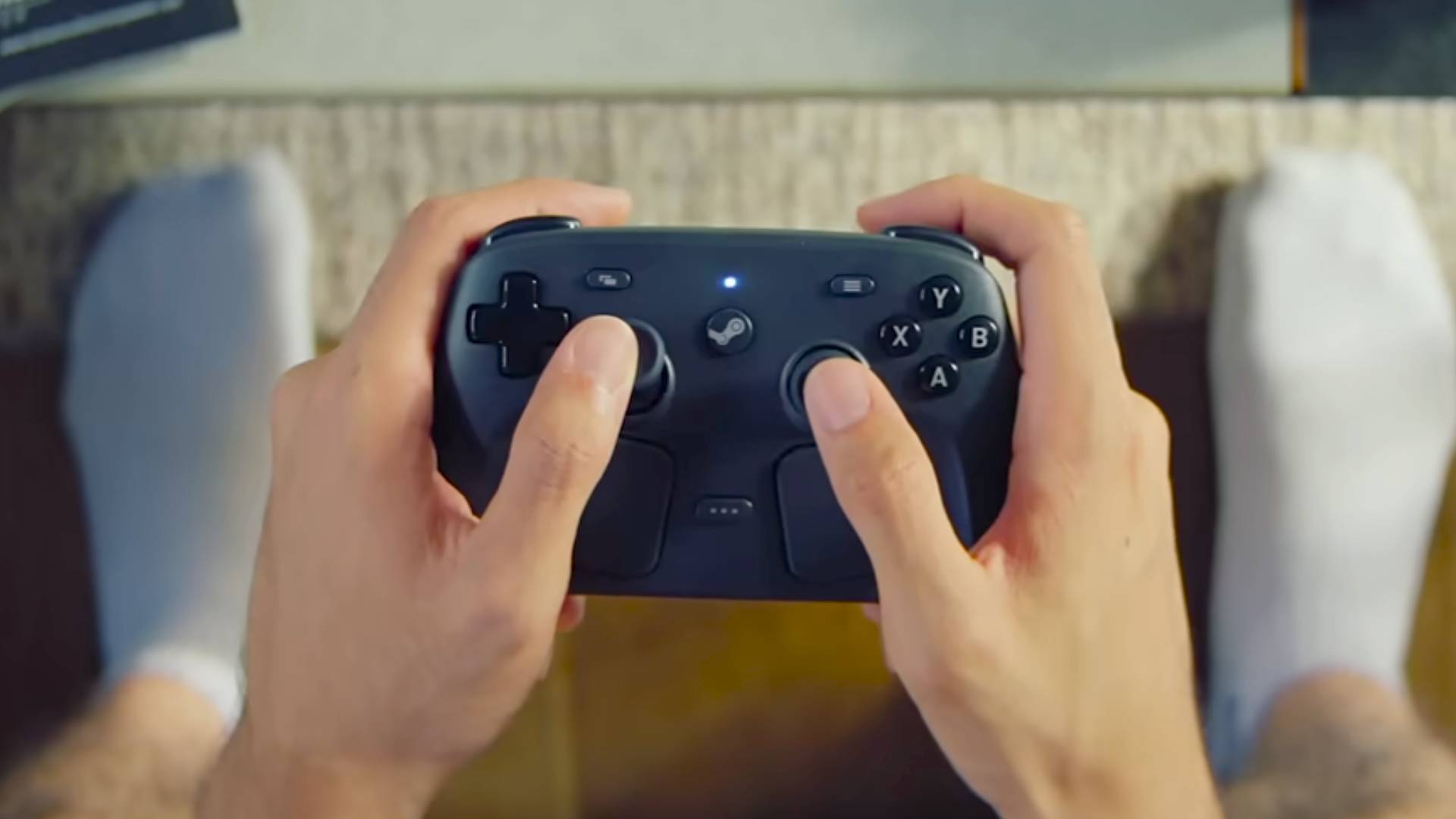 Hands holding Steam Controller with feet wearing socks on floor below.
