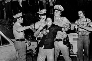 Black and white photograph of a man being dragged away by three police officers, another man stands close by watching the scene