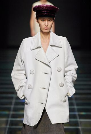 A model from Prada's fall 2024 runway wearing a feather hat with a white peacoat and plaid skirt.