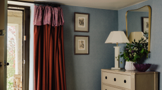 Image of a light blue entryway with the door open and a red curtain by the door. There is a white chest of drawers with a white lamp and flower vase on it and a mirror on the wall.