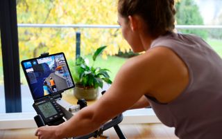 The Concept2 BikeErg is compatible with third party apps via Bluetooth or ANT+ . This image shows the back and right side of a rider looking at a screen showing a virtual ride while on the bike. In the back ground is a plant and balcony.