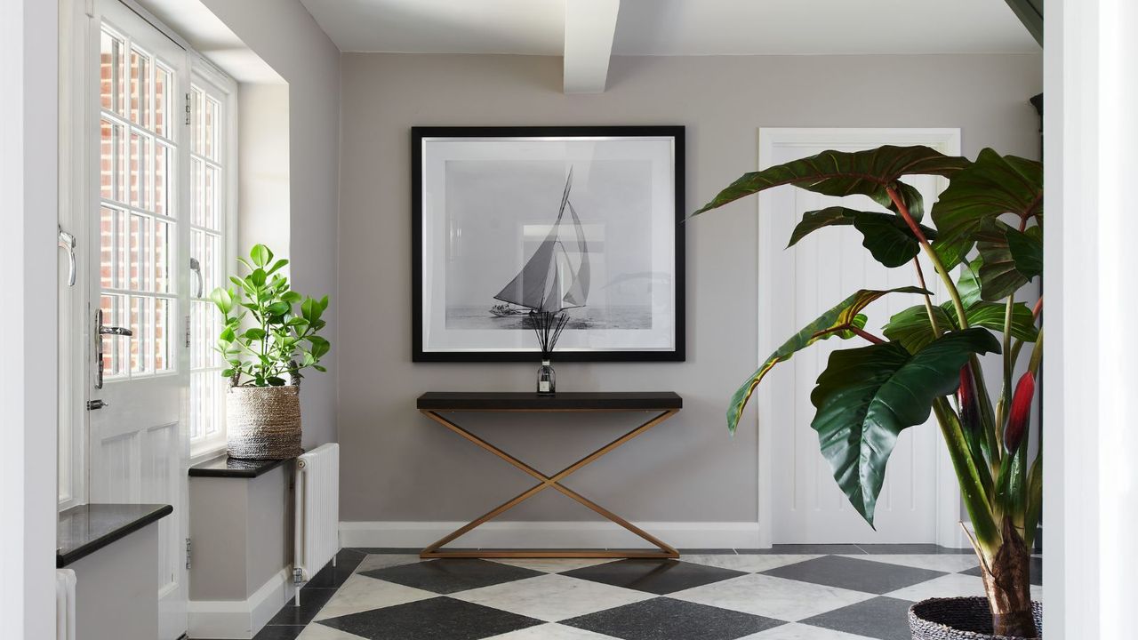An entryway with black and white tiled flooring, a white door to the left, and a black and white framed ship print in the middle with a console table below it