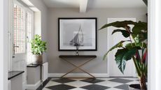 An entryway with black and white tiled flooring, a white door to the left, and a black and white framed ship print in the middle with a console table below it