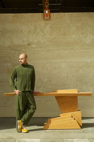 A young man wearing an all-green outfit and yellow shoes stands before a sculptural piece in the tones of ochre in front of a marble wall.